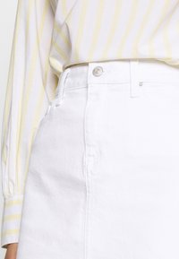 Bílá džínová sukně s vysokým pasem, předními kapsami a stříbrným zapínáním. Nošena s pruhovaným žlutým a bílým topem.