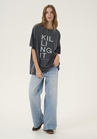 Sivý oversized tričko s nápisom "KILLING IT" v tučných bielych písmenách, skombinované so svetlomodrými širokými nohavicami a hnedými topánkami.