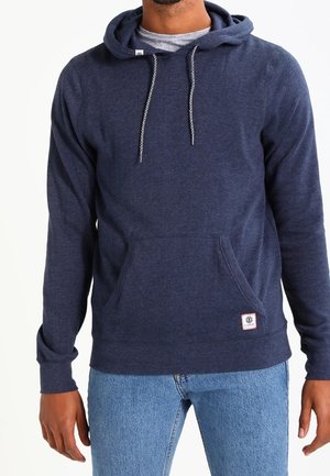 Mand iført en mørkeblå hættetrøje med snore og forlomme, kombineret med lyseblå jeans.