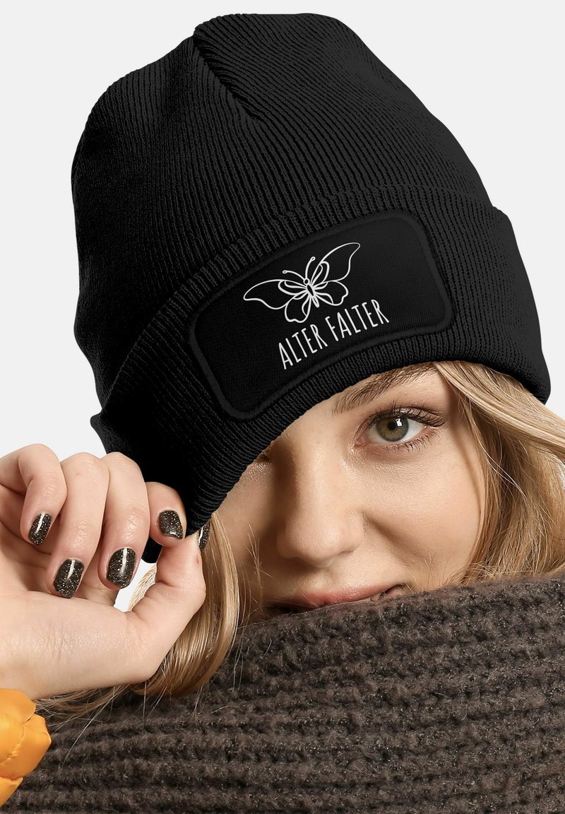 Black ribbed beanie with a rectangular patch featuring a butterfly design and "ALTER FALTER" text, worn while draped in a dark knitted scarf.