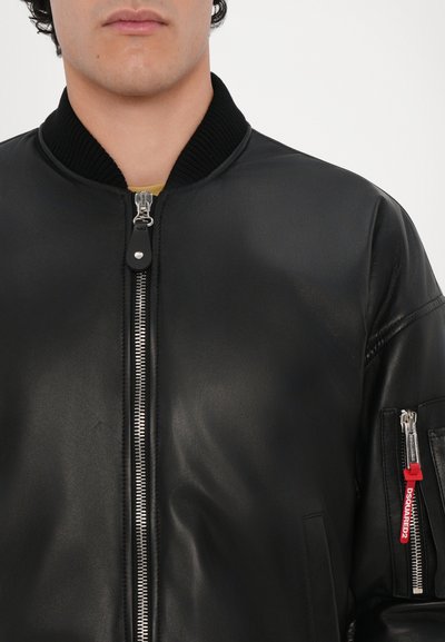 Black leather bomber jacket with a zippered front, ribbed collar, and left sleeve pocket featuring a red accent tag. Smooth texture.
