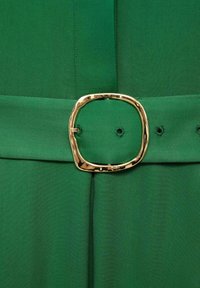 Green fabric with a smooth texture featuring a wide belt. The belt has a chunky, irregular gold buckle and two visible belt loops.