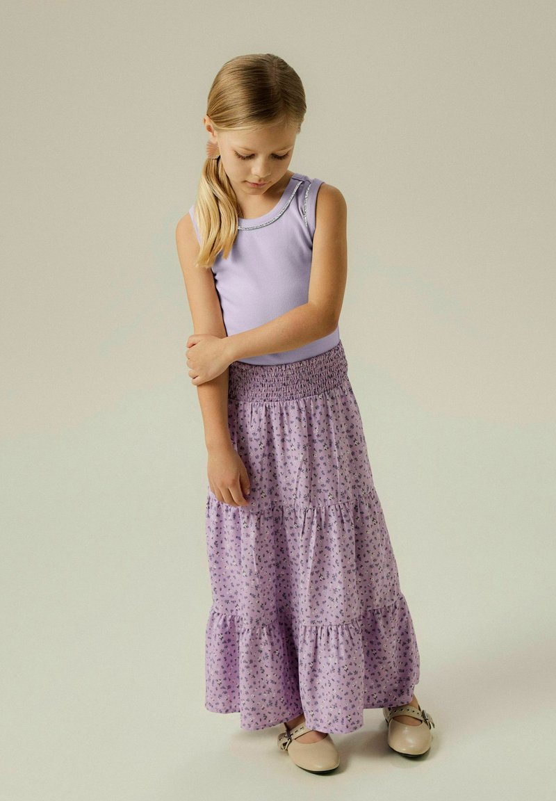 Top de tirantes lila con ribete plateado, combinado con una falda larga de lavanda con estampado floral en capas. Niña con zapatos beige, fondo claro.