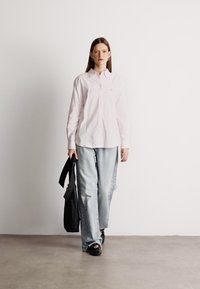 Svijetloružičasta košulja dugih rukava s dugmadi sprijeda, u kombinaciji sa svijetloplavim širokim trapericama. Crna torba preko ramena držana u ruci. Jednostavna pozadina.