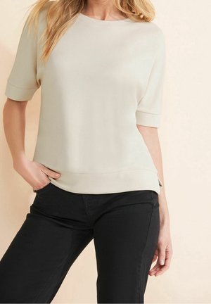 Femme portant un haut crème à manches courtes avec des revers aux manches et un pantalon noir, debout avec une main dans la poche et l'autre détendue.
