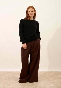 Schwarzer strukturierter Pullover kombiniert mit weit geschnittenen braunen Hosen. Das Outfit zeichnet sich durch eine lässige Passform und gerippte Säume aus und wird vor einem neutralen Hintergrund präsentiert.