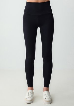 Leggings neri a vita alta realizzati in tessuto elasticizzato, con una texture liscia e gambe affusolate, abbinati a scarpe da ginnastica bianche.