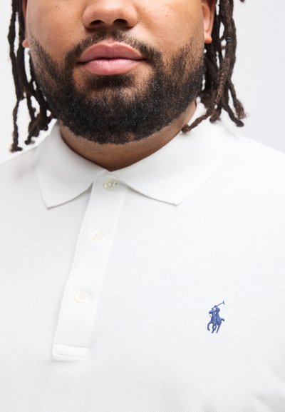 Άνδρας με γενειάδα και dreadlocks που φοράει λευκό πόλο πουκάμισο με μικρό μπλε κεντημένο λογότυπο παίκτη πόλο στο στήθος.