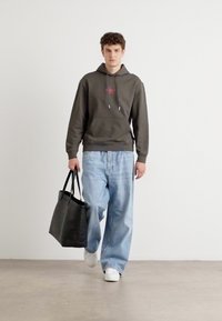 Borsa a tracolla nera in tela, felpa grigia con logo rosso, jeans larghi in denim blu e sneakers bianche, su un modello che cammina davanti a uno sfondo semplice.