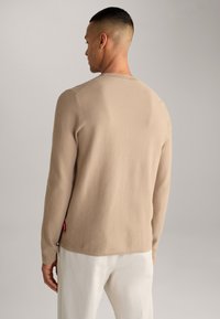 Pull en tricot côtelé beige, à manches longues, avec un col rond et une petite étiquette rouge sur le côté, associé à un pantalon de couleur claire.