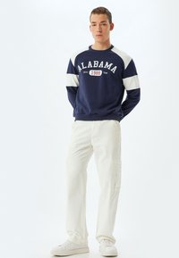 Marineblaues Sweatshirt mit weißen Raglanärmeln, Text "ALABAMA 1988", kombiniert mit weißen Cargo-Hosen und weißen Sneakers, stehend auf einem neutralen Hintergrund.