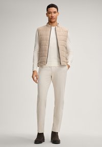 Beige gesteppte Weste, darunter ein cremfarbenes Sweatshirt, helle Hose und dunkle Stiefeletten. Einfaches Design und glatte Texturen.