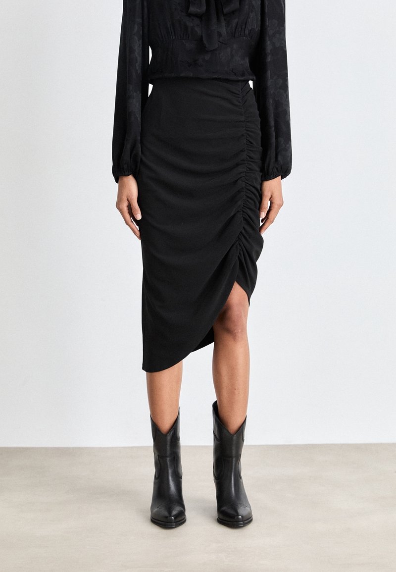 Femme portant une jupe asymétrique noire froncée, un chemisier noir à manches longues et des bottines noires sur un fond clair uni.