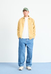 Gul skjorta över en vit t-shirt, blå vida jeans, grön keps och ljusa sneakers. Enkel design utan synliga mönster.