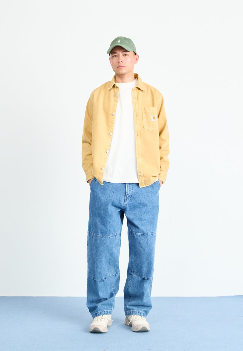 Gul skjorta över en vit t-shirt, blå vida jeans, grön keps och ljusa sneakers. Enkel design utan synliga mönster.