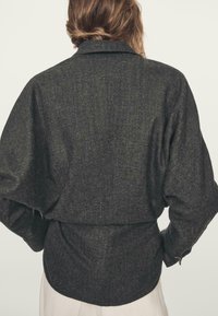 Ciemnoszara koszula z mieszanki wełny o luźnym kroju, z kołnierzykiem typu fold-over, długimi rękawami oraz detalem w formie ściągnięcia w pasie, z subtelną teksturą.