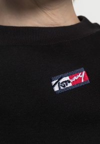 Acercamiento a una camisa de tela negra con relieve, que presenta un pequeño parche bordado rectangular en rojo, azul y blanco con texto parcialmente visible.