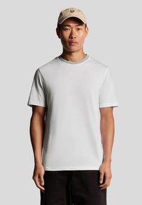 T-shirt bianco a maniche corte con collo rotondo, abbinato a un cappellino beige con un piccolo logo, indossato su uno sfondo grigio.