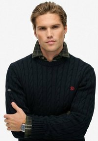 Junger Mann mit verschränkten Armen, der einen dunklen, strukturgewebten Pullover über einem grünen Karoshemd trägt, mit einer am linken Handgelenk sichtbaren Armbanduhr.