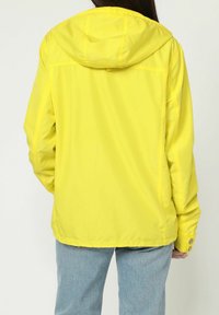 Chaqueta amarilla fabricada con un material ligero e impermeable, con capucha, mangas largas y detalles de botones a presión. Textura suave, corte casual.