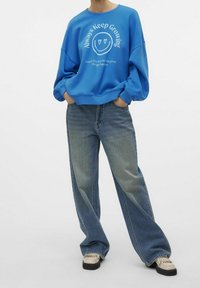 Sudadera azul con texto gráfico blanco y cara sonriente, combinada con vaqueros de pierna ancha de color azul claro y zapatos beige con acentos.