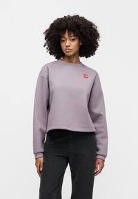 Ανοιχτό μωβ cropped φούτερ με στρογγυλή λαιμόκοψη και ριμπ τελείωμα στα μανίκια, με μικρό κόκκινο λογότυπο στο αριστερό στήθος. Συνδυάζεται με μαύρο παντελόνι.