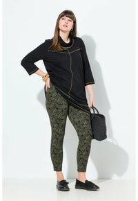 Donna che indossa una tunica nera a maniche lunghe con cuciture gialle, leggings con motivo verde e nero, mocassini neri e tiene una borsa a tracolla nera intrecciata.