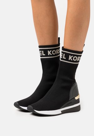 Botines de punto negros con un diseño elástico, que cuentan con una banda de logo blanca en la parte superior y una suela de cuña con un detalle dorado.