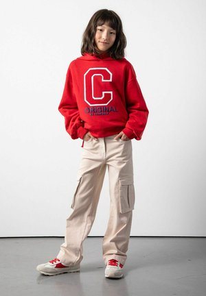 Czerwona bluza z kapturem z dużą białą literą "C" i niebieskim napisem "ORIGINAL", w połączeniu z beżowymi spodniami cargo z bocznymi kieszeniami i sneakersami z czerwonymi akcentami.