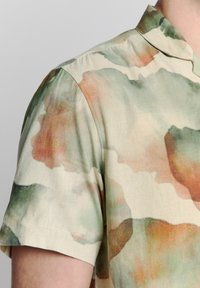 Hemd met korte mouwen van lichte stof, met een aquarelpatroon in groene en oranje tinten op een crèmekleurige achtergrond.