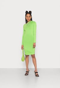 Vestido justo de manga longa verde com detalhe franzido, combinado com uma mala verde correspondente e sandálias pretas de salto alto. Textura suave.