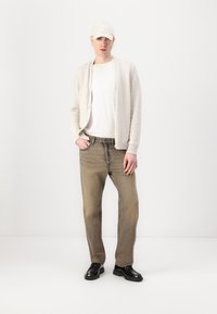 Beige knit cardigan, white t-shirt, faded light brown jeans, black shoes, and a light cap; casual fit with button accents and a relaxed silhouette.