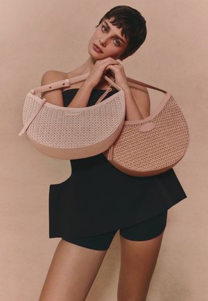 Donna con vestito nero che tiene due grandi borse intrecciate a forma di mezzaluna, una beige e una marrone chiaro, su uno sfondo neutro.