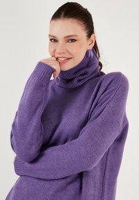 Vijolični pulover z dolgimi rokavi iz mehkega materiala, z rebrastimi manšeti in sproščenim krojem. Podrobnosti vključujejo udoben, zvit ovratnik.