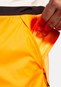 Shorts orange avec une poche latérale zippée ; texture lisse ; ceinture noire ; main tirant un objet plié de la poche.
