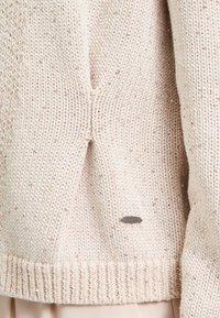 Beige knitted sweater featuring a textured pattern, decorative metallic threads, and a small branded label near the hem.