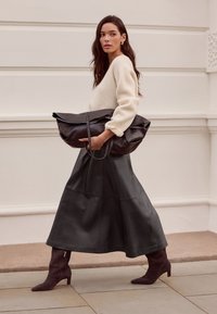 Schwarze Ledertasche mit großer Überklappklappe, getragen zu einem cremefarbenen, gerippten Pullover und einem dunkelbraunen Lederrock in A-Linie, ergänzt durch violette Knöchelstiefel.