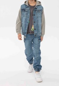 Chaleco de denim con capucha gris, cierres de botón y dos bolsillos frontales, combinado con pantalones de denim azul sueltos con puños elásticos y bolsillos tipo cargo.
