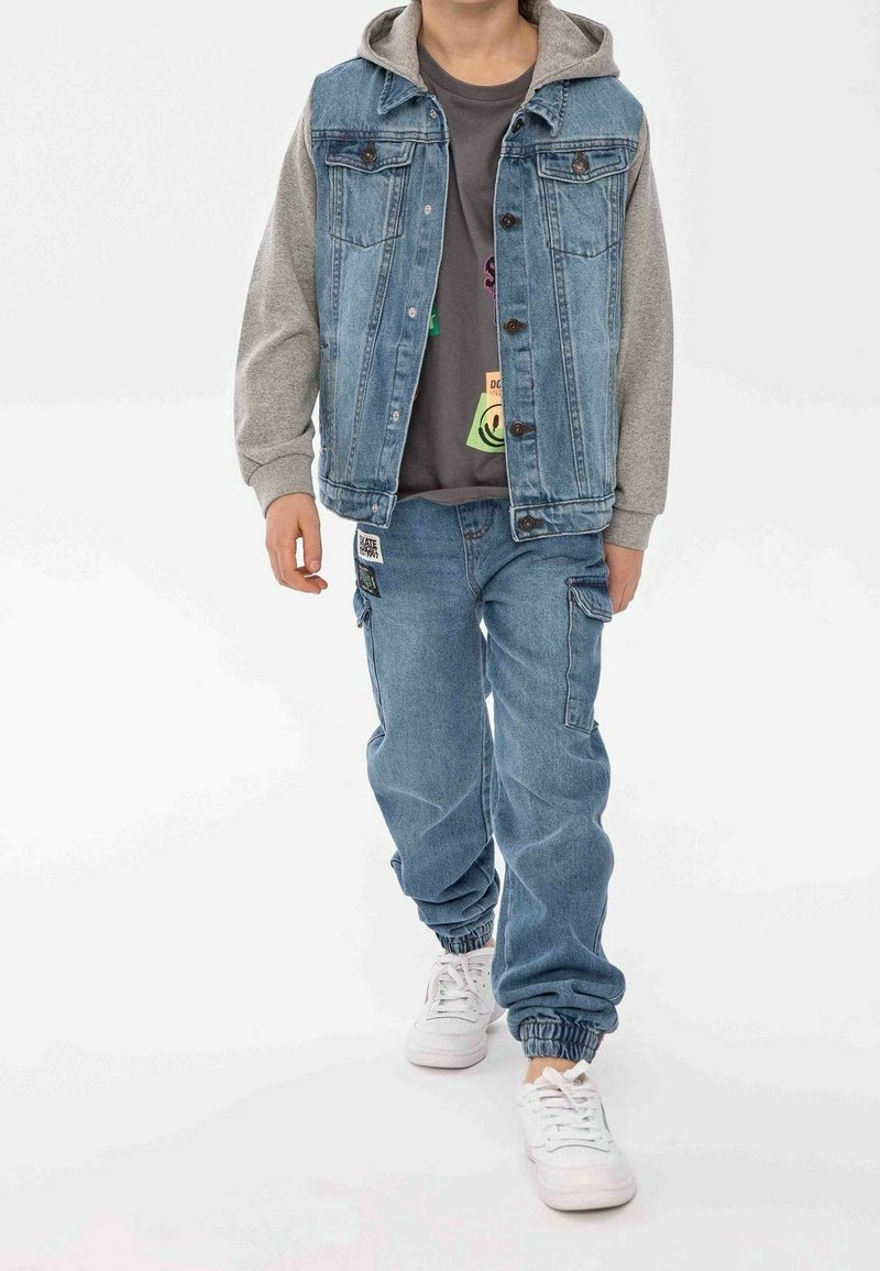 Chaleco de denim con capucha gris, cierres de botón y dos bolsillos frontales, combinado con pantalones de denim azul sueltos con puños elásticos y bolsillos tipo cargo.