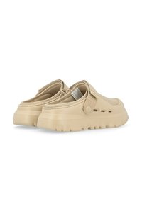 Zapatillas de color beige sin cordones con un upper suave y texturizado, suela elevadora y una correa lateral. El diseño incluye orificios de ventilación y suela estriada.