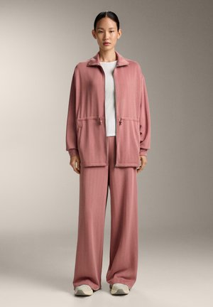 Femeie în picioare, purtând o jachetă roz deschisă, largă, cu fermoar și pantaloni largi asortați, cu adidași albi și bluză albă, pe un fundal neutru.