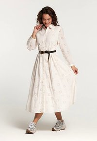 Lange witte jurk met een bloemrijk ajourpatroon, knopen aan de voorkant, kraag en elastische taille, gecombineerd met een zwarte riem en grijze sportschoenen.