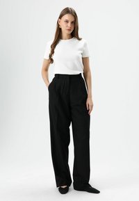 Weißes T-Shirt mit kurzen Ärmeln kombiniert mit schwarzen, hoch-taillierten, weit geschnittenen Hosen. Das Model trägt schwarze spitze Ballerinas. Glatte Textur, minimalistisches Design.