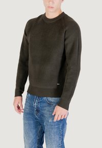 Uomo che indossa un maglione a coste verde scuro e jeans blu sbiaditi, in piedi contro uno sfondo chiaro uniforme.