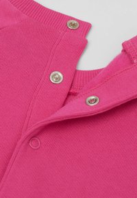 Růžový pletený oděv s límcem, se kovovými patentkami a texturovaným povrchem. Tkanina je měkká a hladká, zdůrazňující podrobné švy.