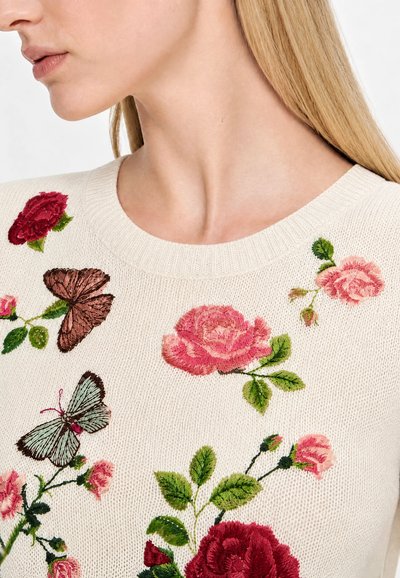 Nærbillede af en kvinde iført en cremefarvet sweater broderet med lyserøde og røde roser og to sommerfugle på bryst- og skulderområdet.