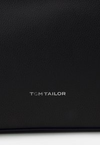 Černý kožený produkt s texturovaným povrchem. Obsahuje stříbrné logo "TOM TAILOR" na spodní straně, což naznačuje identitu značky.