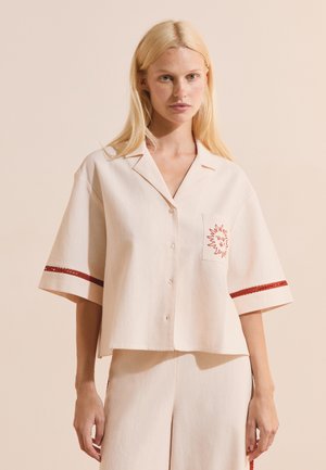 Jeune femme aux longs cheveux blonds portant une chemise crème ample à manches courtes avec une broderie rouge de soleil et un pantalon large assorti.