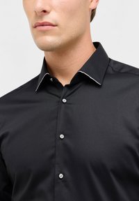 Zwarte button-up shirt met een gestructureerde stof, contrasterende witte accenten op de kraag, en vijf knopen aan de voorkant.