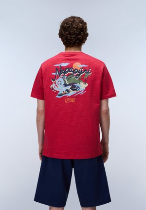 Mand iført rød T-shirt med farverigt tropisk strandmotiv og teksten "Napapijri Est 1987," kombineret med marineblå short, set bagfra.